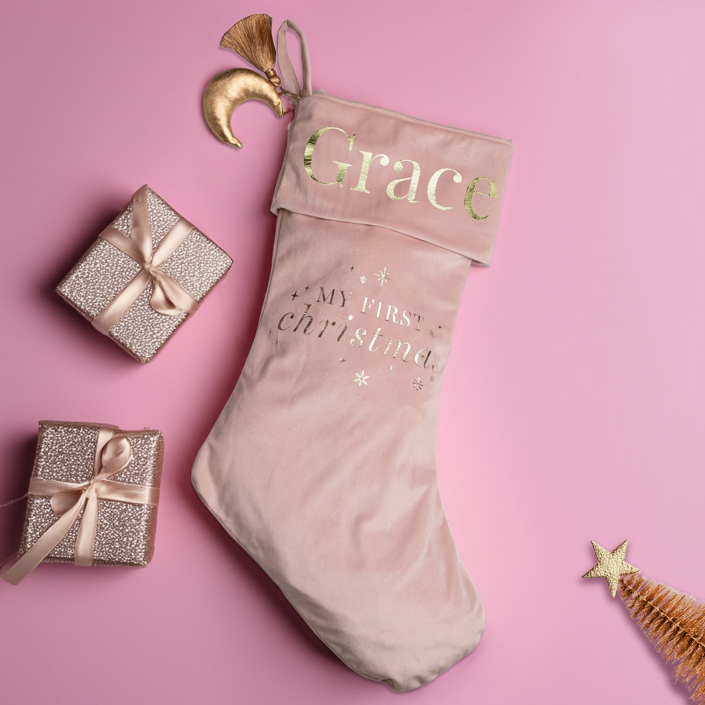 Personalised my First Christmas stocking surrounded by decorative items on a pink background.