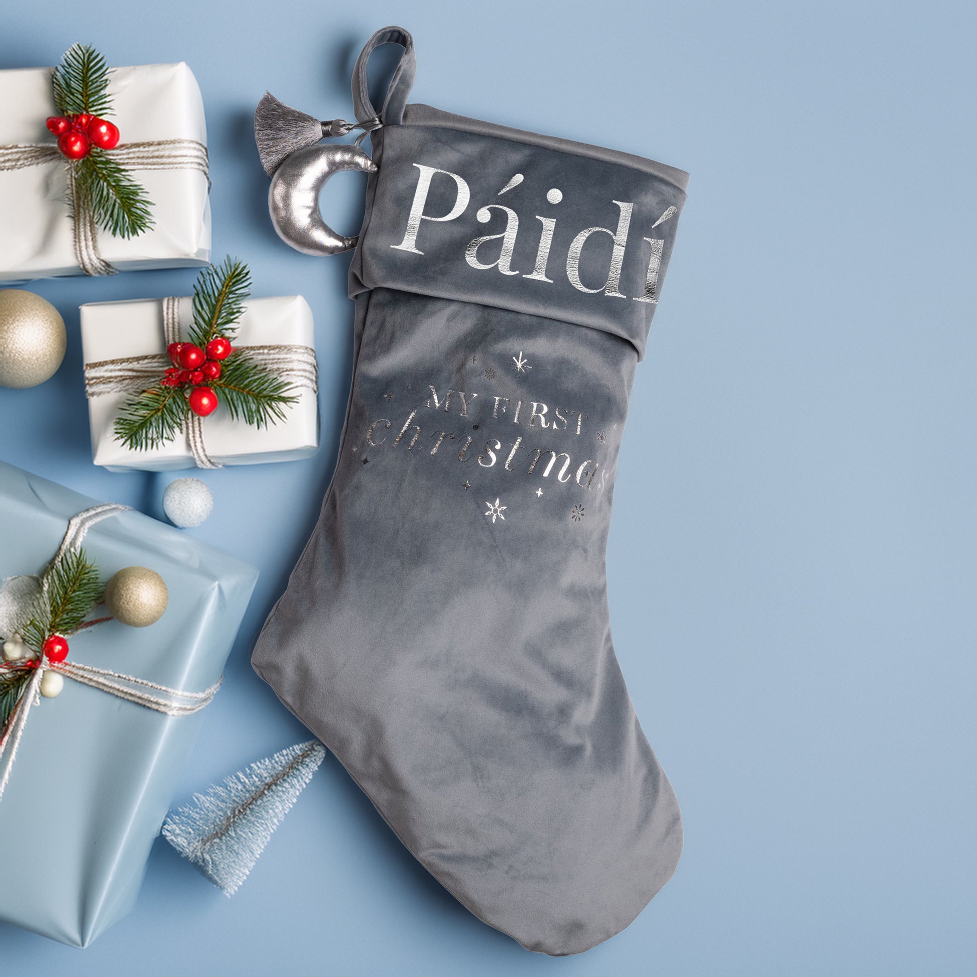 Personalised my First Christmas stocking surrounded by decorative items on a blue background.